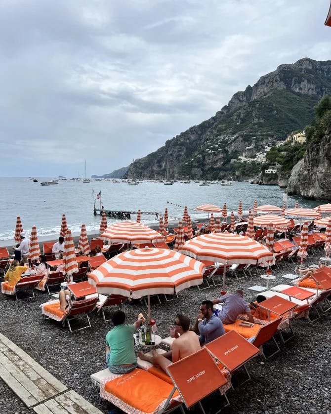A picture of people enjoying at Arienzo Beach in Italy surrounded by orange lawn chairs and orange and white striped umbrellas with the mountain and ocean in the background