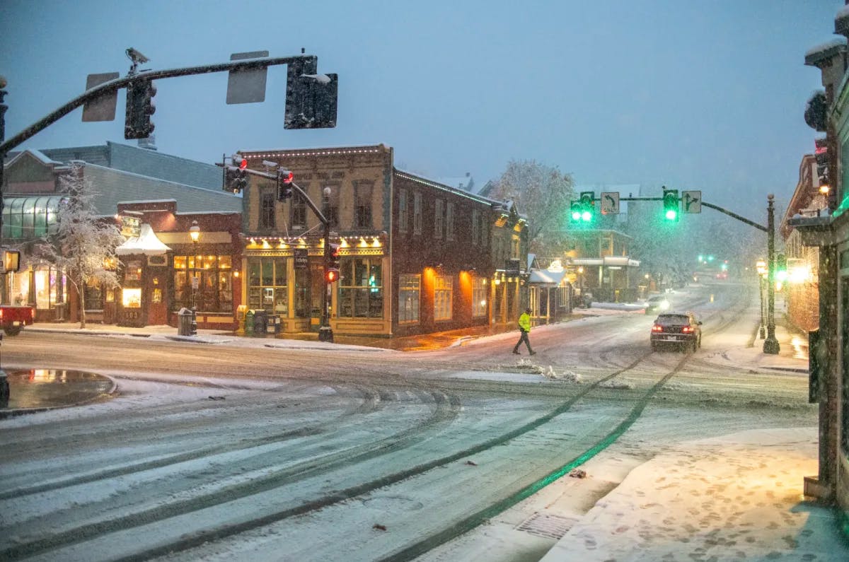 A view of a snowy main street with buildings, green street lights, cars, a pedestrian walking and exterior lights turned on. 
