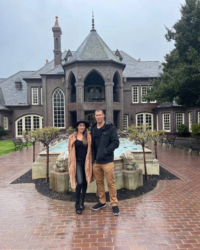Elyssa and her partner posing outside of a mansion in front of a landscaped pond while standing on a brick driveway