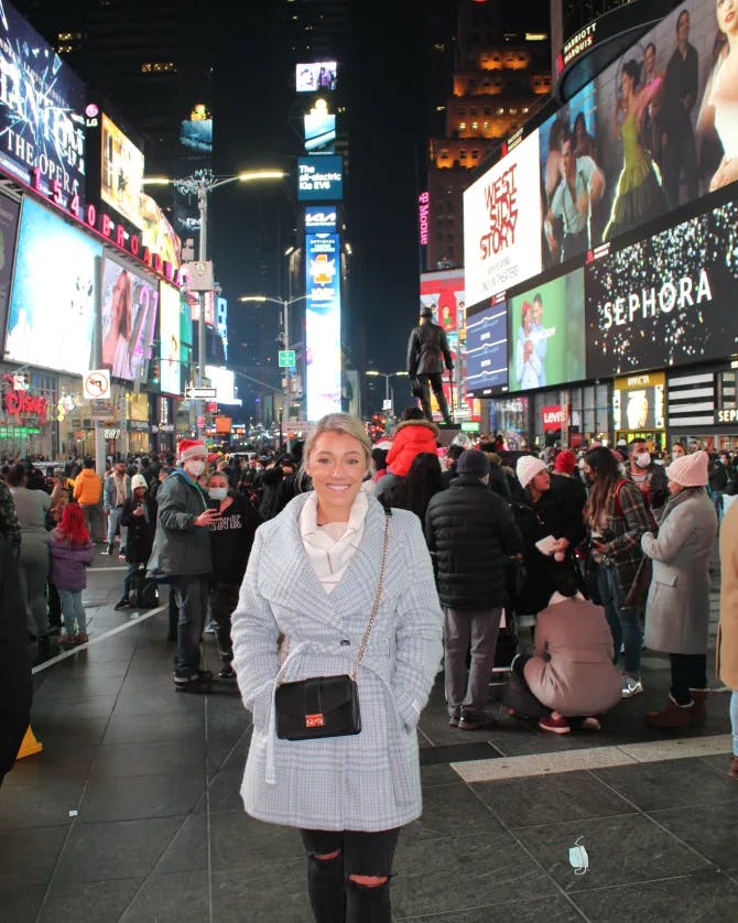 Zoe wearing a light blue coat, black pants and bag while posing in front of Times Square in New York City