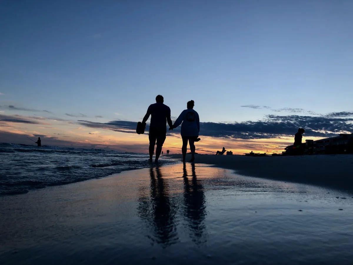 A picture of a couple walking on the shore during the sunset.