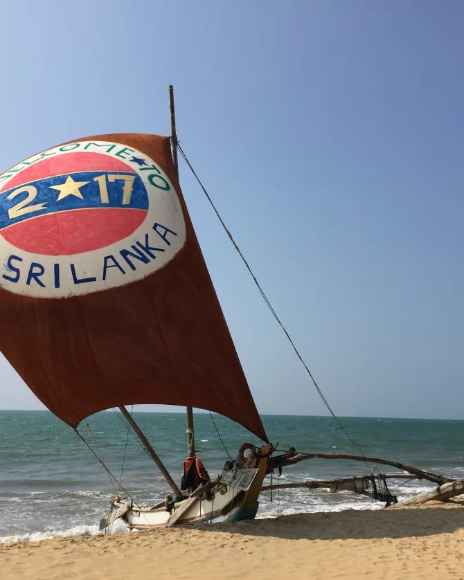 A boat with a large red sail anchored on a beach with the sea in the background