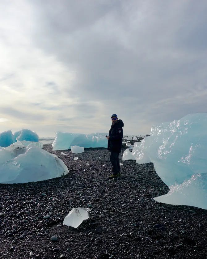 Kara standing on a rocky shore with large ice bergs around her and a shoreline to the left
