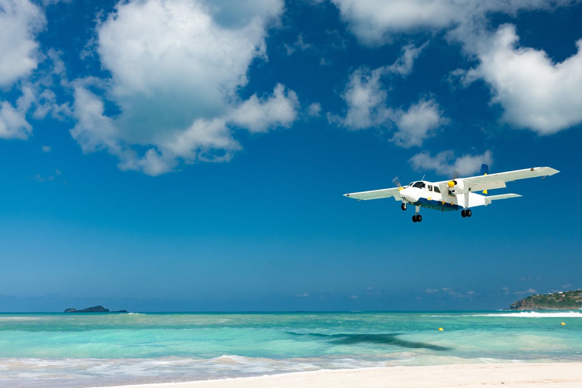 Small white plane flies over white sand beach and turquoise waters on a sunny day in St. Barths