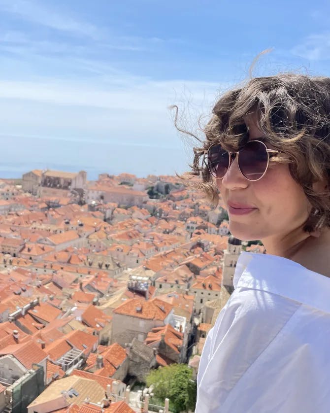 Emily wearing a white top and sunglasses while posing in front of an aerial view of red-roofed buildings and the blue sea in the background.