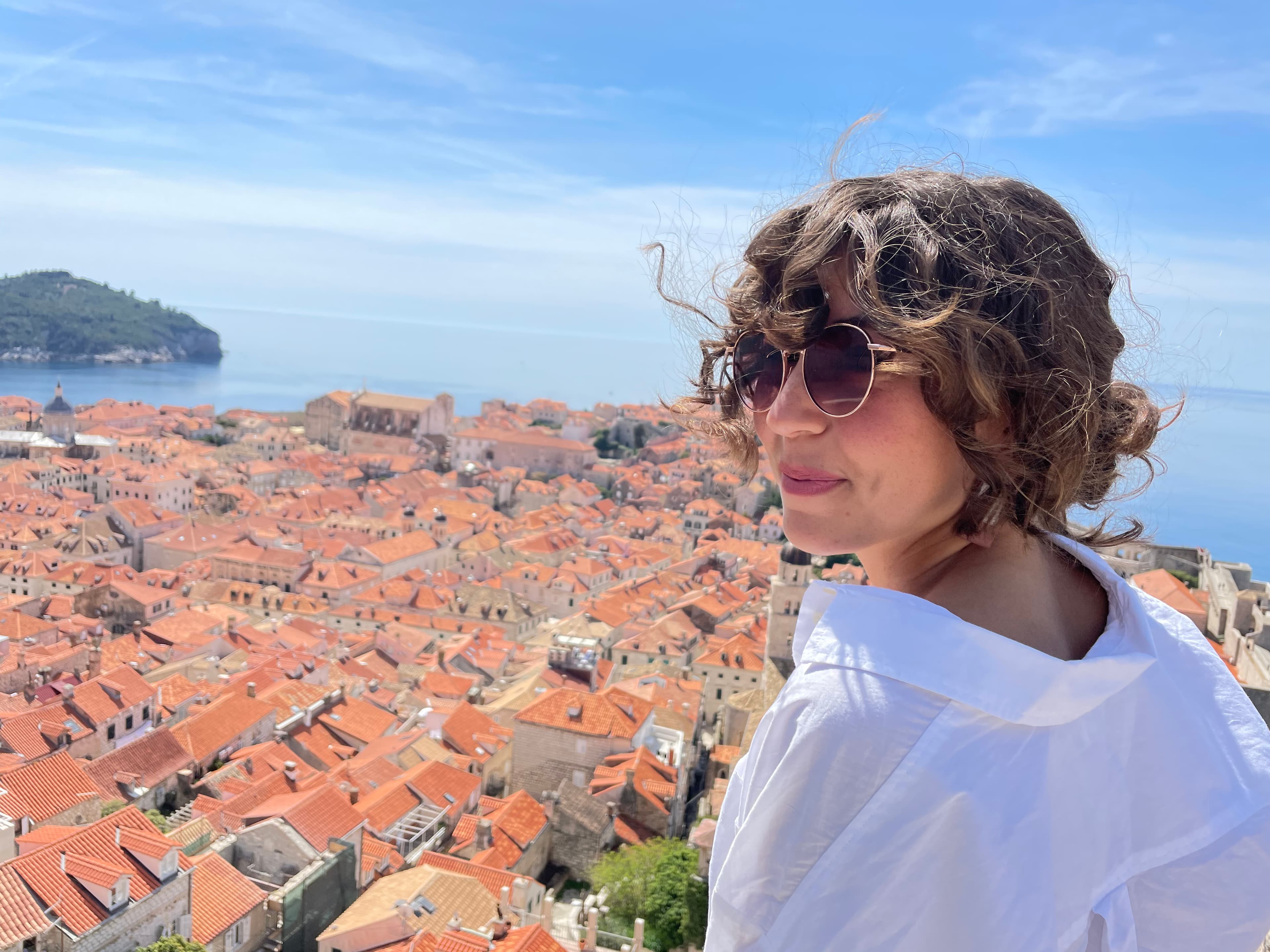 Emily wearing a white top and sunglasses while posing in front of an aerial view of red-roofed buildings and the blue sea in the background.