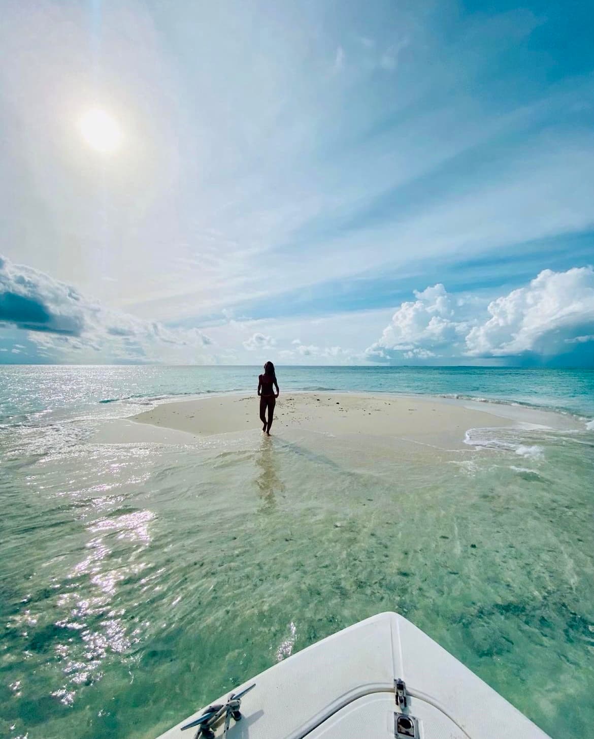 A picture of a female silhouette walking forward on a sand bank surrounded by blue water. The sun is shining bright from above surrounded by blue sky and clouds.
