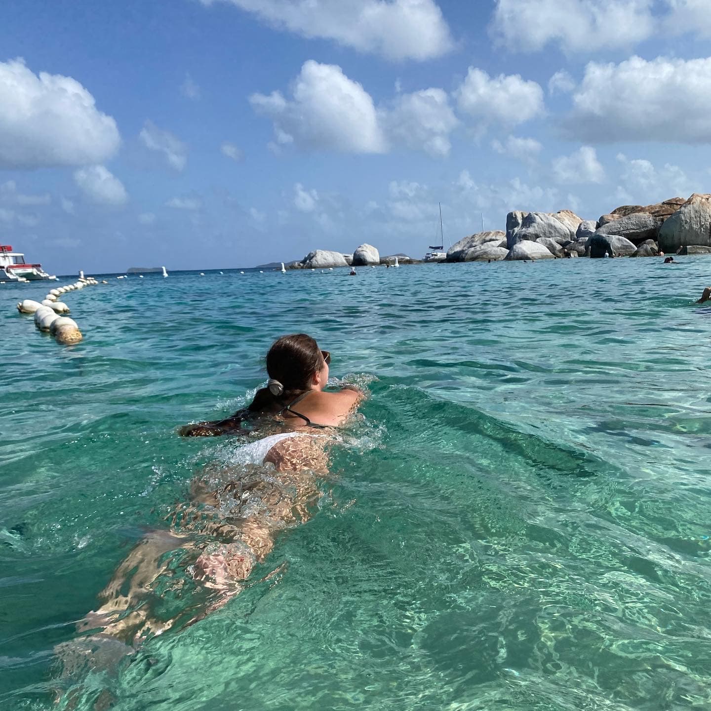 Samantha swimming in clear turquoise waters towards a jetty of large rocks.