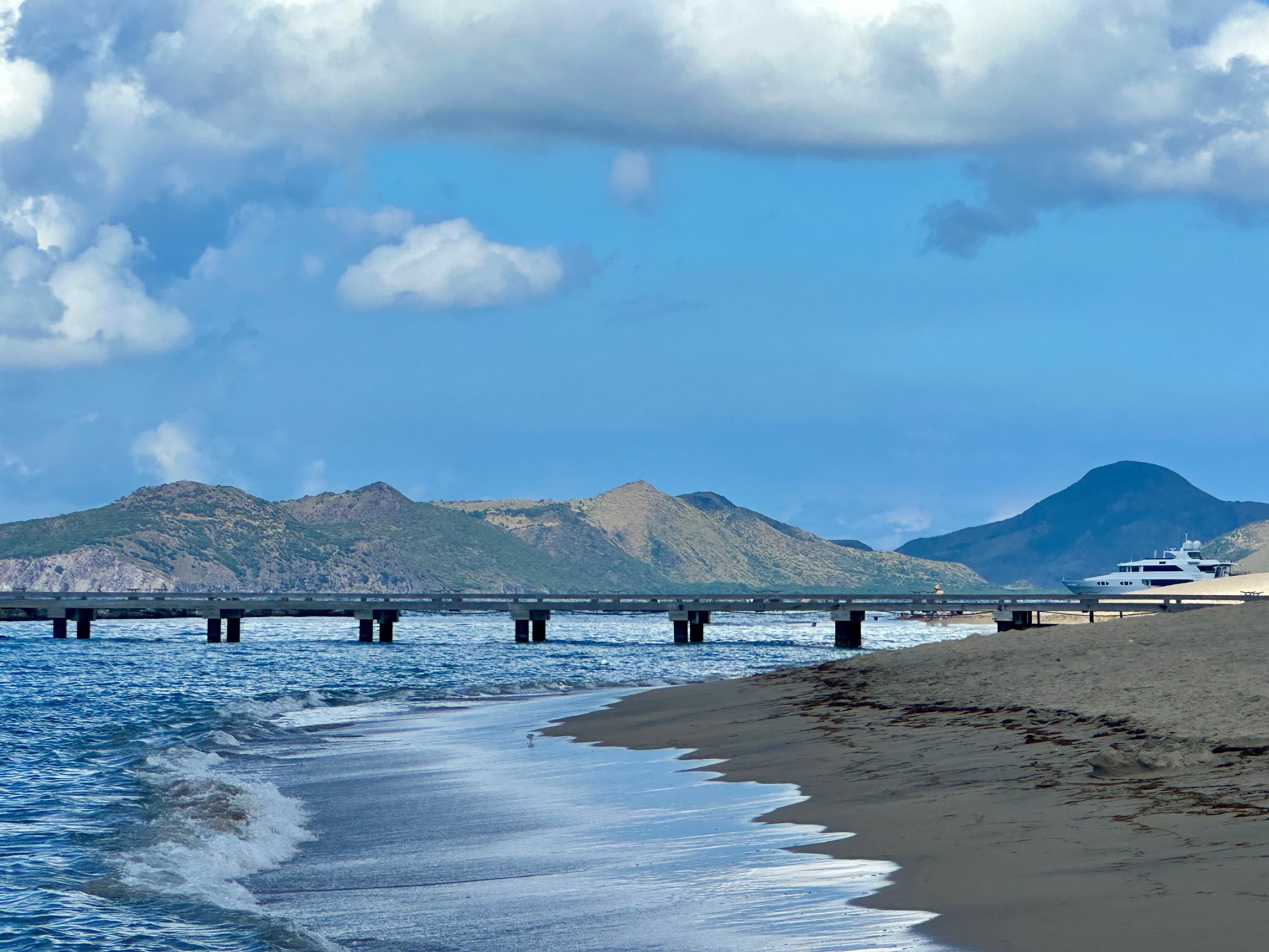 Photo of the seashore with a jetty, ship, and mountains in the background.