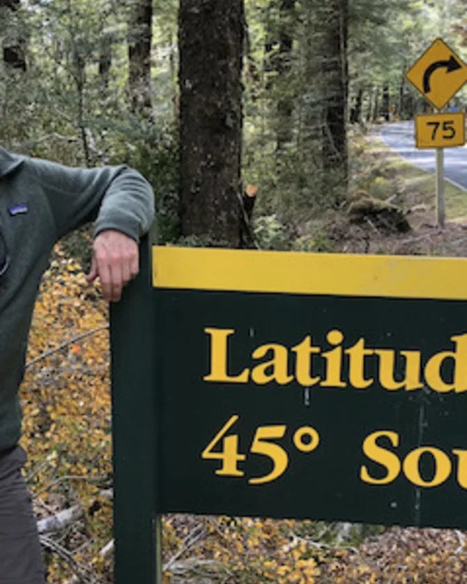 Ken standing in front of a road sign that reads 'Latitude 45º South' in a forest setting