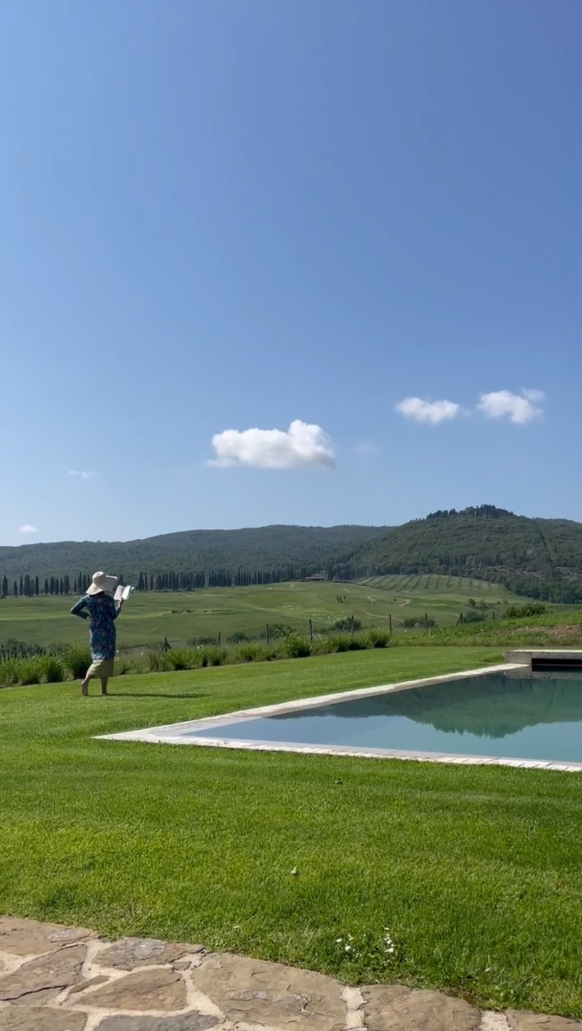 Kiki reading a book while walking along the grass surrounding a swimming pool with mountains in the background