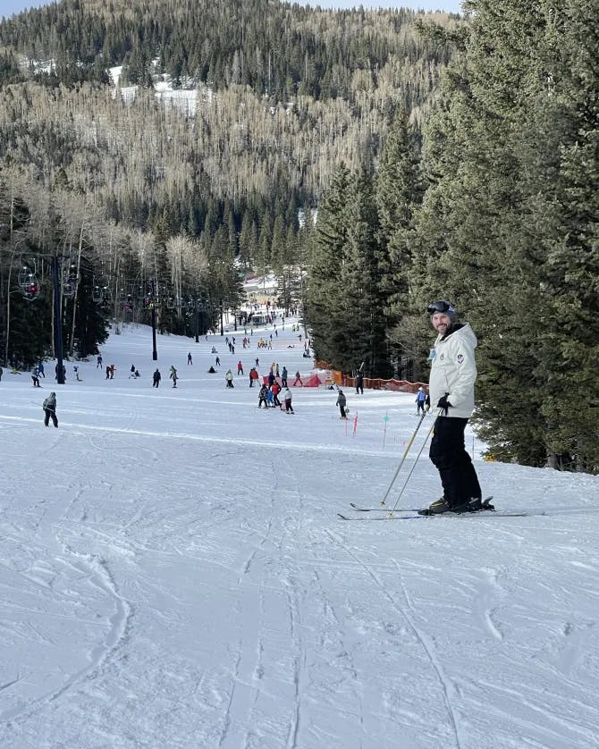 Vanessa wearing a white coat and black pants while posing on a ski hill surrounded by snowy pine trees.