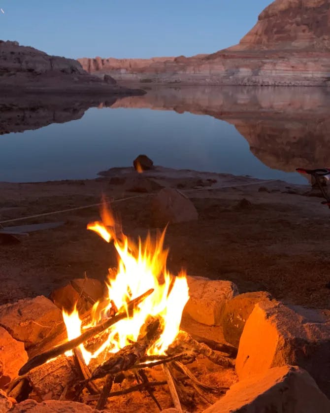 A campfire on a beach near a lake with red rocks in the background.