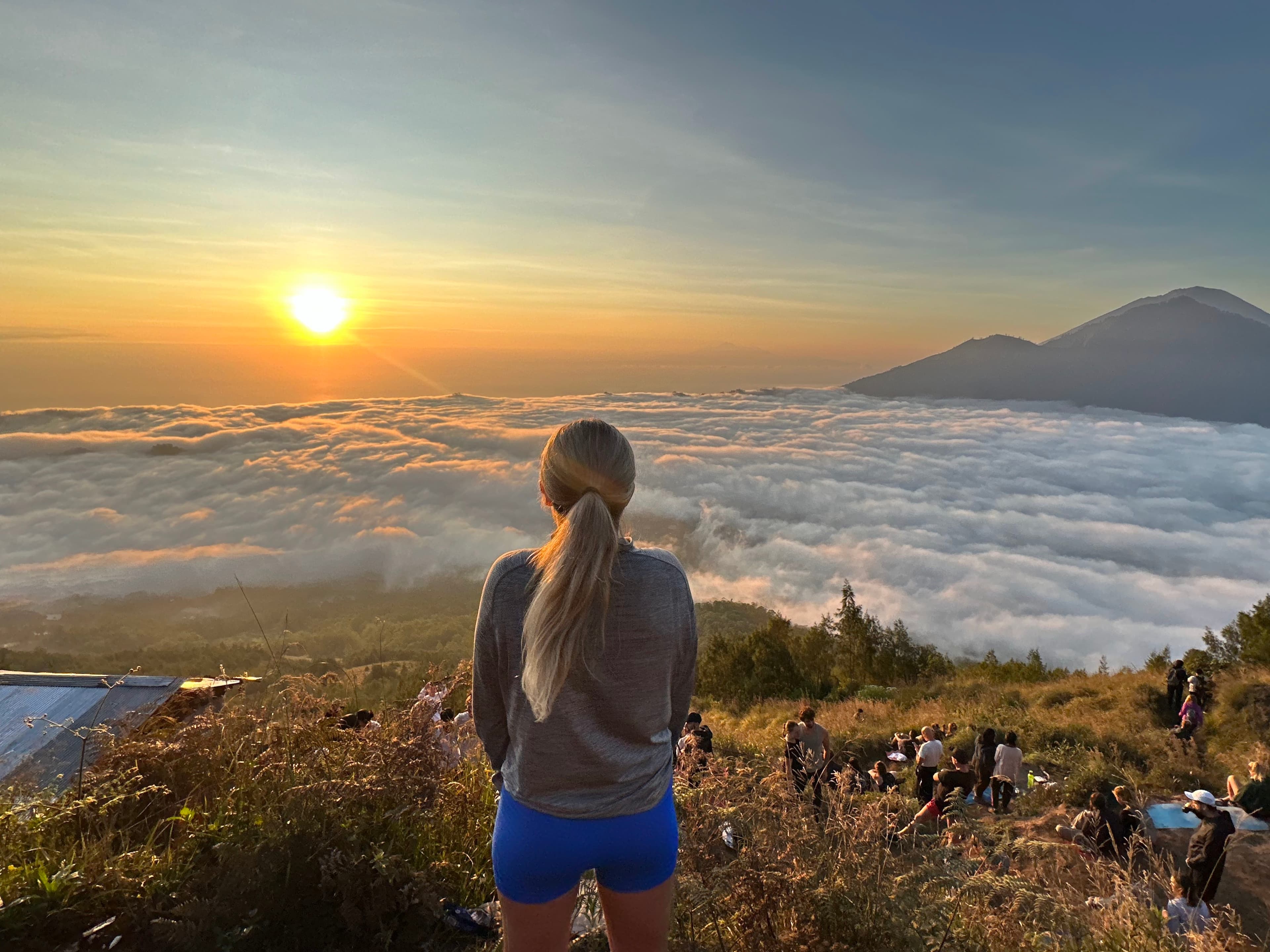 Carly stands above the clouds on top of a mountain with the view of the rising sun and the peak of another mountain in the distance.