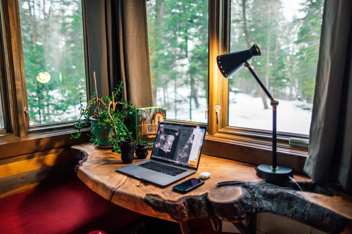 A rustic-theme home office. Through cabin windows: snow is on the ground while evergreen trees still possess lush leaves