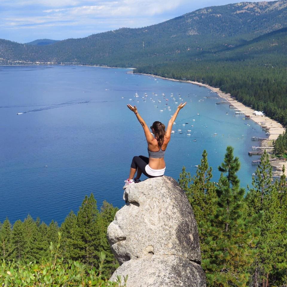 Angela sitting on Monkey Rock with her hands reaching above her head looking out onto an ocean view surrounded by green mountains
