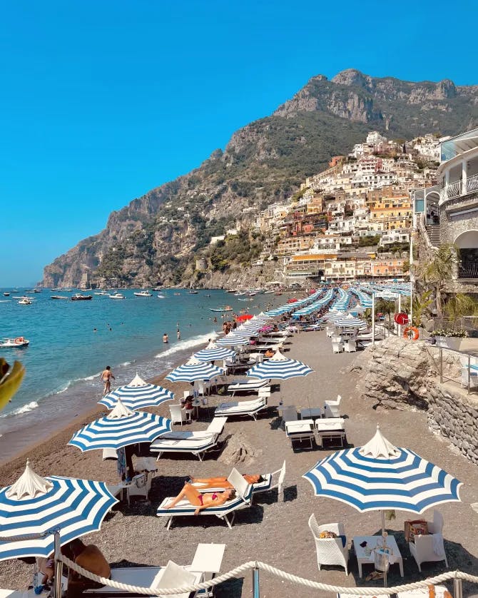 Picture of Amalfi Coast beach with blue and white striped umbrellas lined up in front of a cascade of colorful buildings set into a mountain overlooking the water