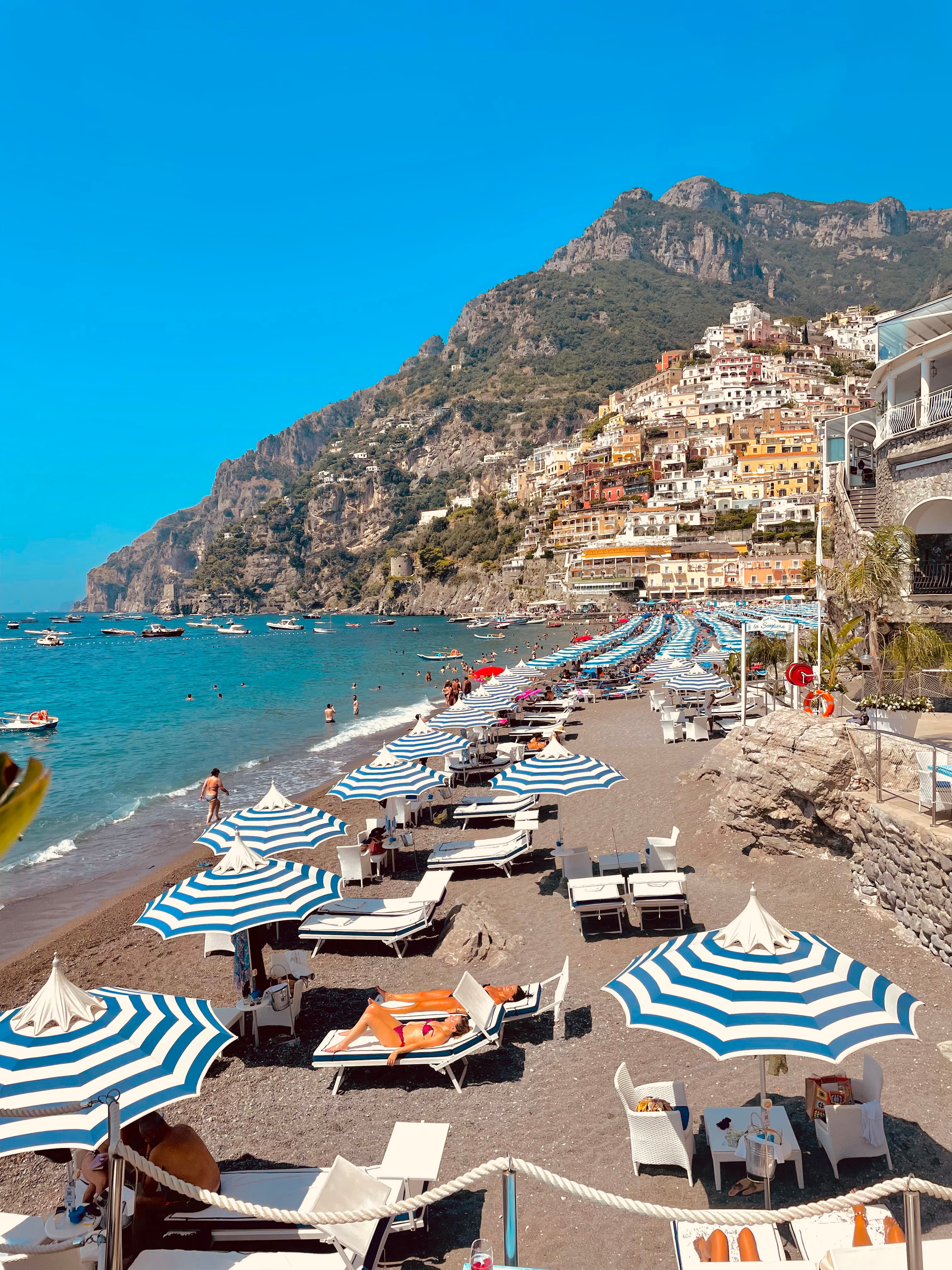 Picture of Amalfi Coast beach with blue and white striped umbrellas lined up in front of a cascade of colorful buildings set into a mountain overlooking the water