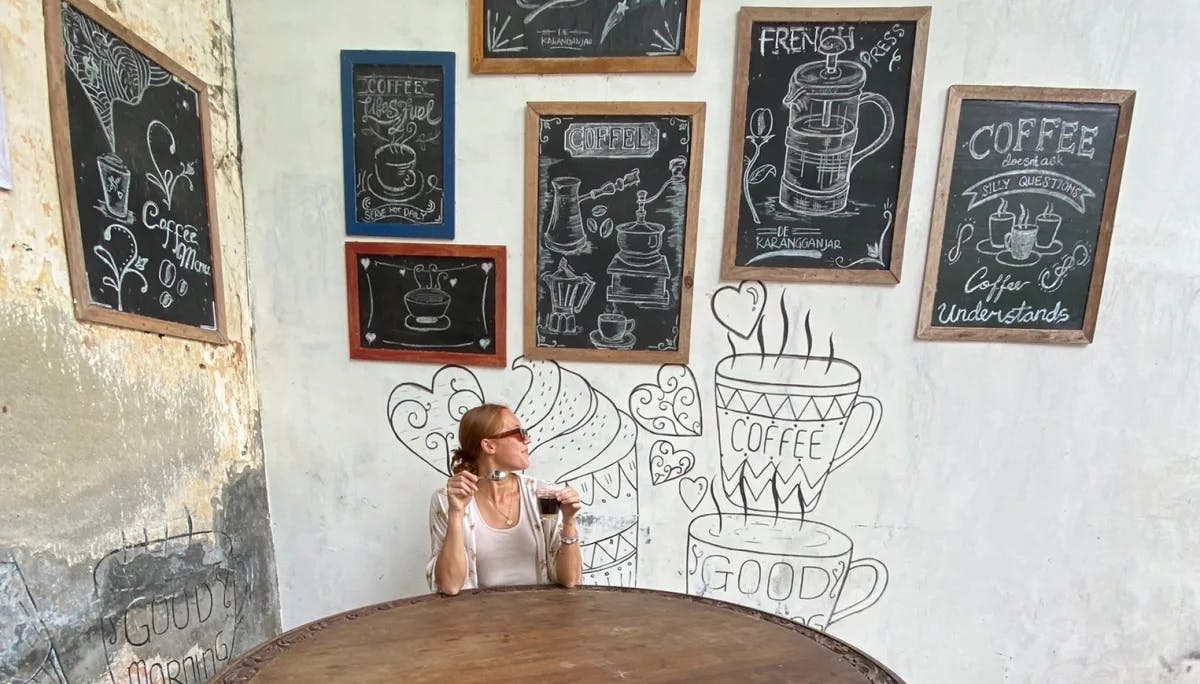 Isabel sitting at a cafe behind a wooden table and in front of a wall with chalkboards, art and a drink in hand.