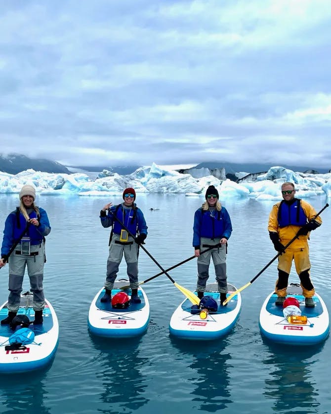 Four people paddle boarding on water with glaciers and dark clouds in the background