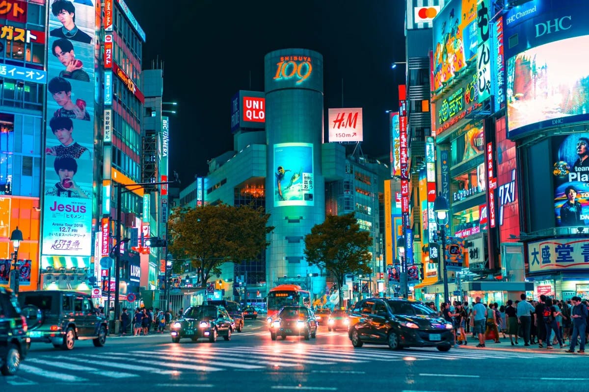 Neon billboards light up a busy intersection in Shibuya, Japan as hundreds of locals stroll along the sidewalks and cars pass through