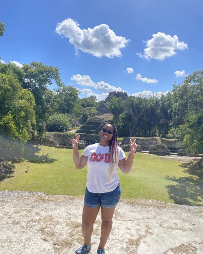 Jia wearing a white top and denim shorts posing with peace sign hand signals while standing on a dirt path in front of green grass and trees on a sunny day 