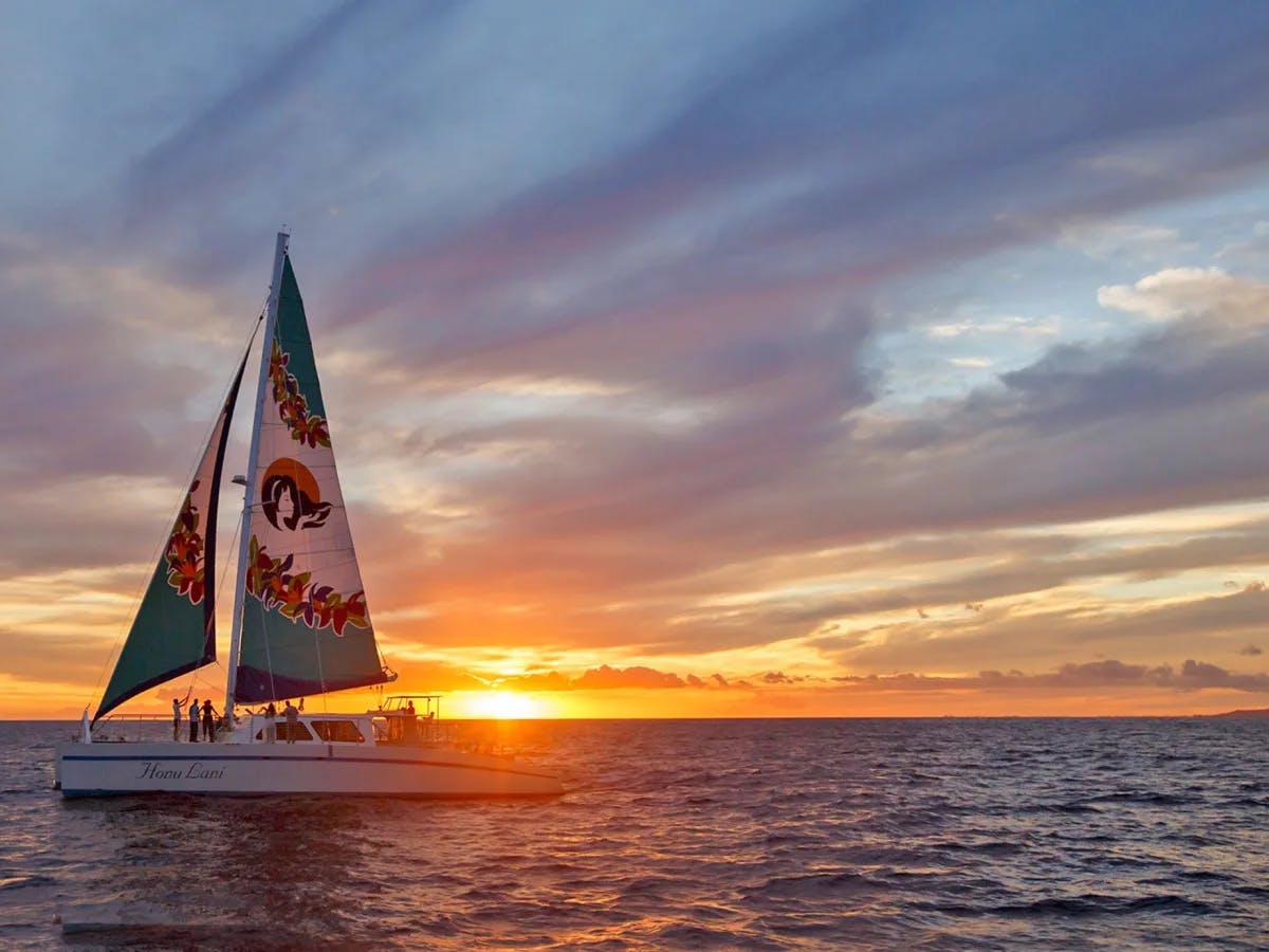 The Hanu Lani sailboat, with locally decorated sails, on the water at sunset.