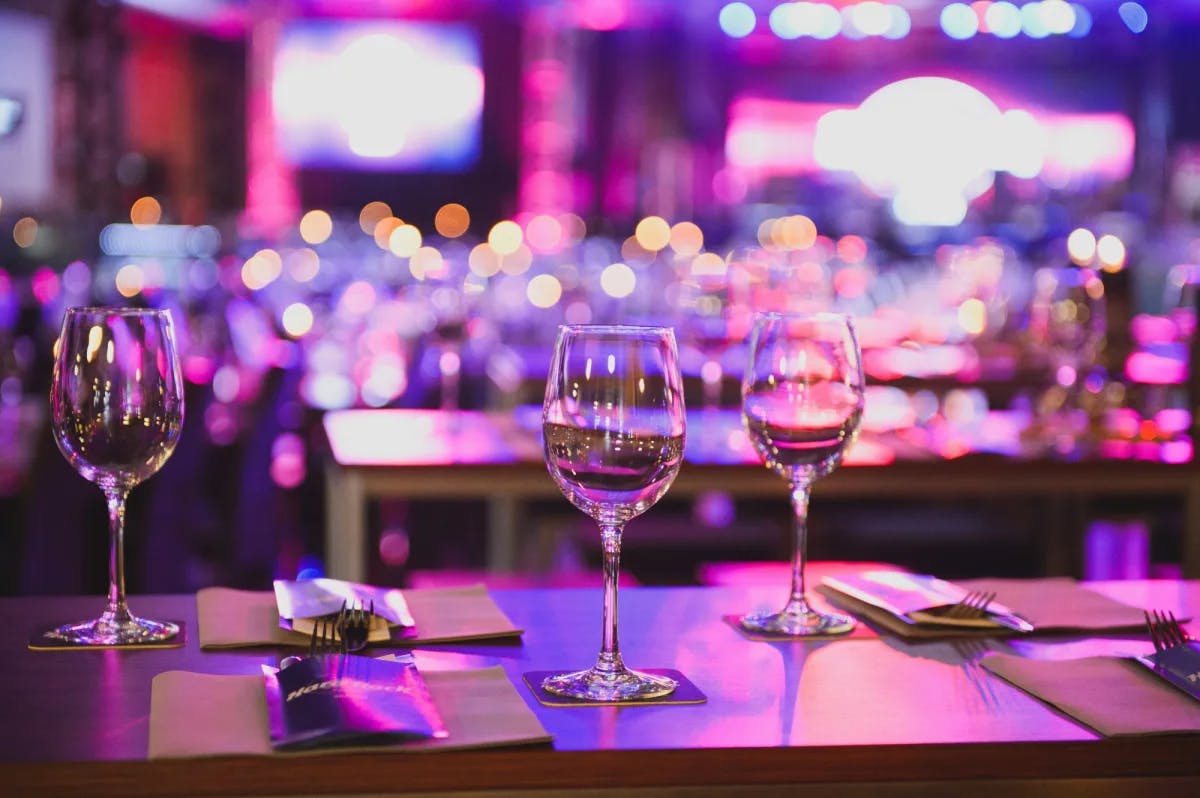 Three wine glasses and table settings in the forefront of an interior image of a restaurant with bright purple lighting blurred out in the background. 