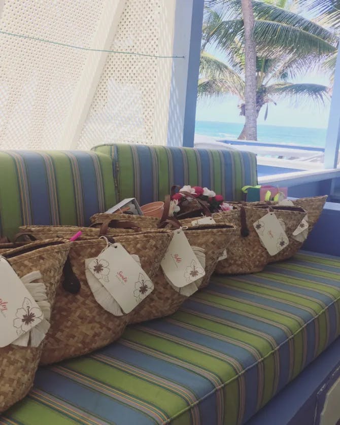 A blue and green striped couch with straw bags placed on top of it and a palm tree and ocean in the background