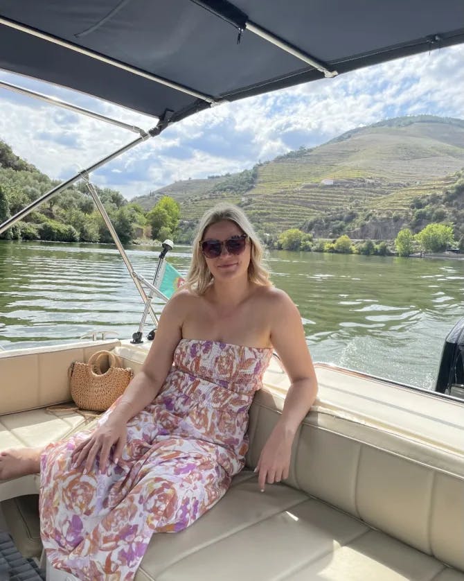 Picture of Ashley sitting on a boat, wearing a colorful dress with the water and mountain in the background