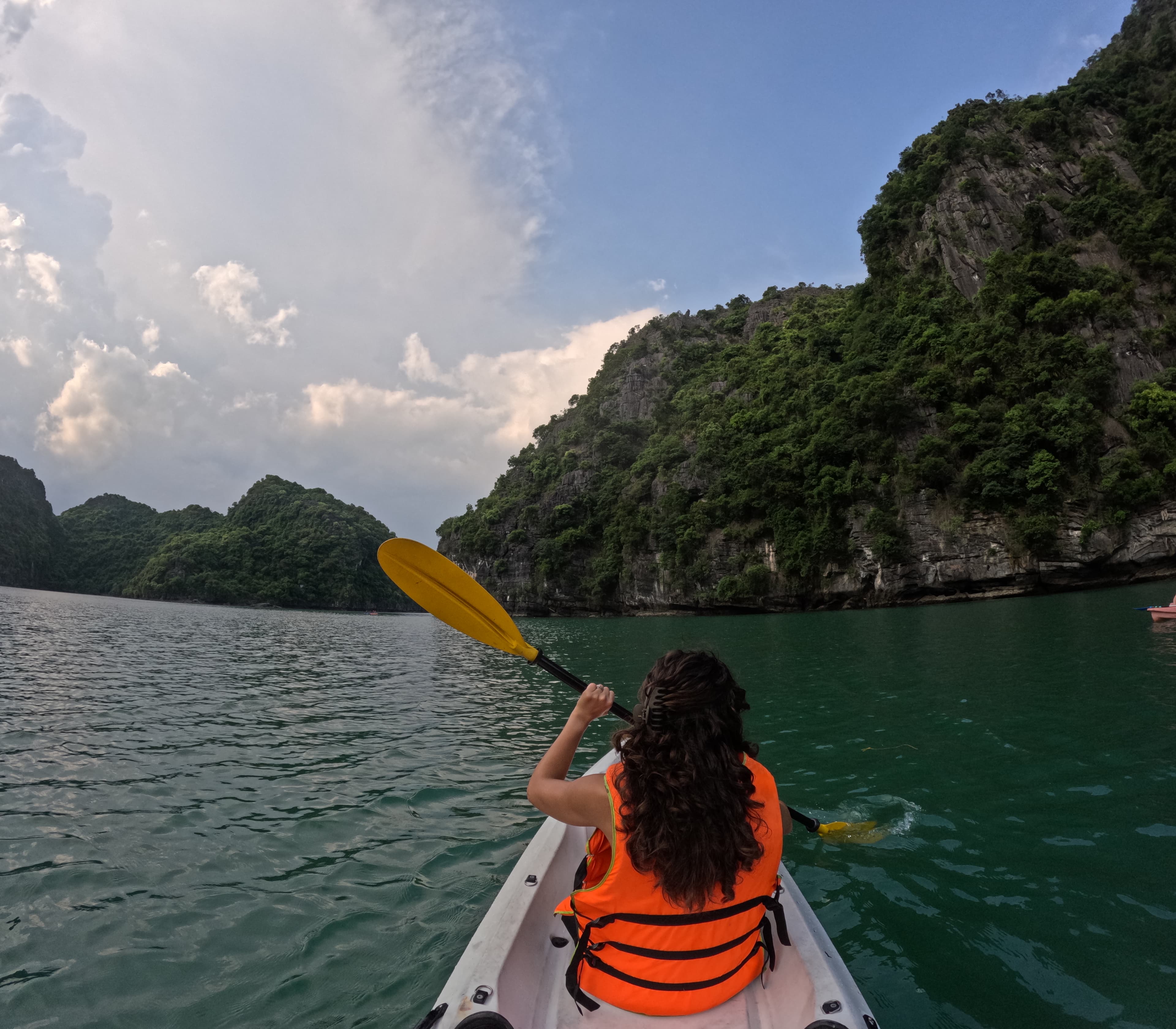 Emily wearing an orange life-vest and kayaking on a body of water near green mountains.