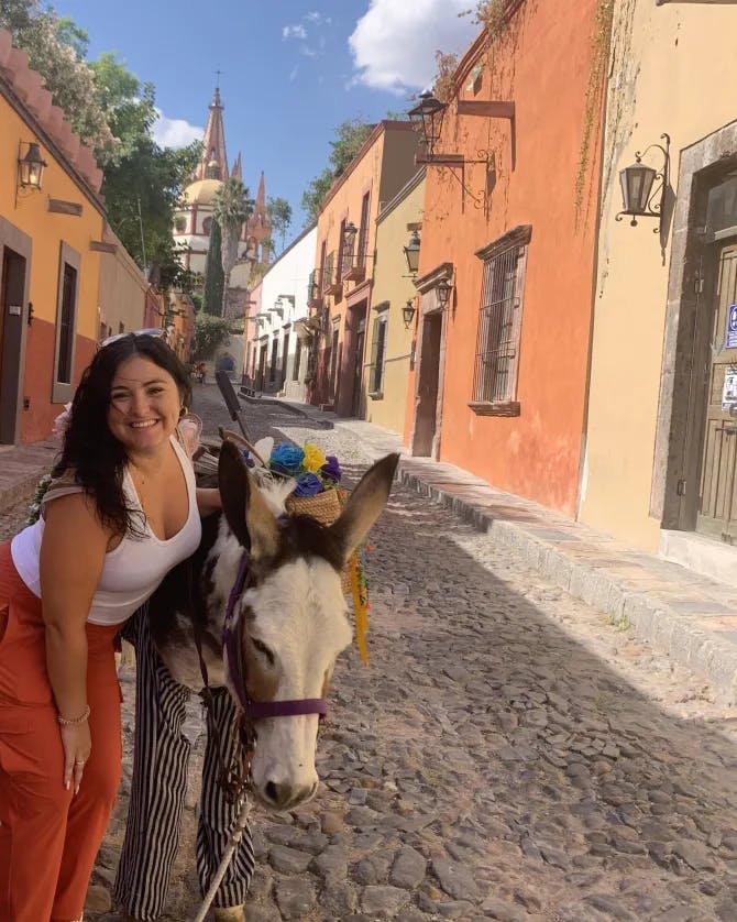 Picture of Brittany wearing a white shirt and red pants leaning down next to a Mule in the street surrounded by colorful buildings 