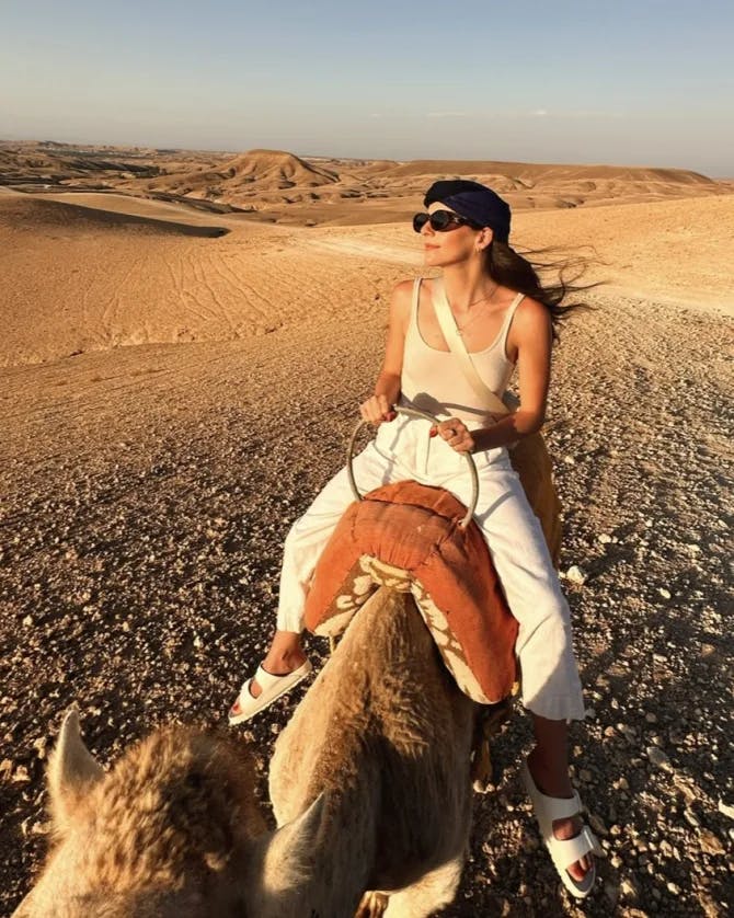 Gabby wearing a white outfit and sunglasses riding a camel in the desert