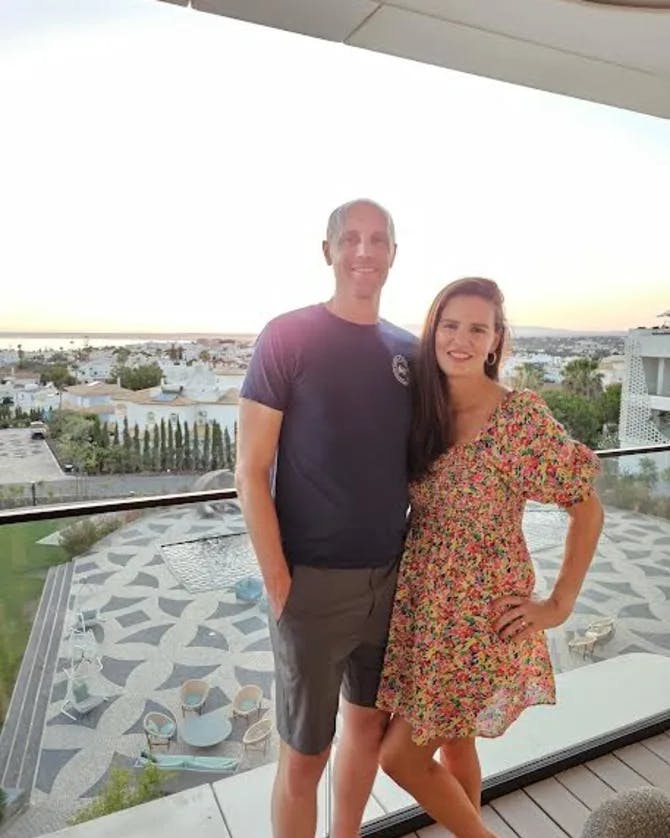 A picture of Meredith wearing a floral dress and posing with her spouse in front of a patio view at sunset