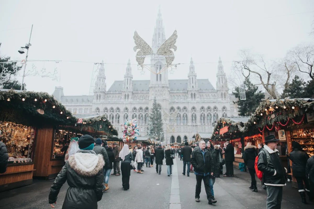 Travelers and locals peruse a Christmas market in Vienna while majestic Old World architecture stands prominently in the foggy background