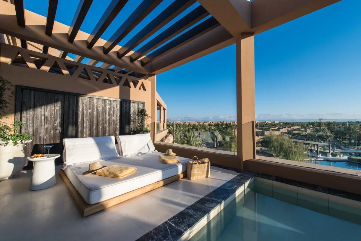 Foreground: a two-person daybed sits atop a rooftop terrace with a cocktail sitting on a nightstand and infinity pool. Background: the grounds of the resort with the Atlas Mountains in the far distance