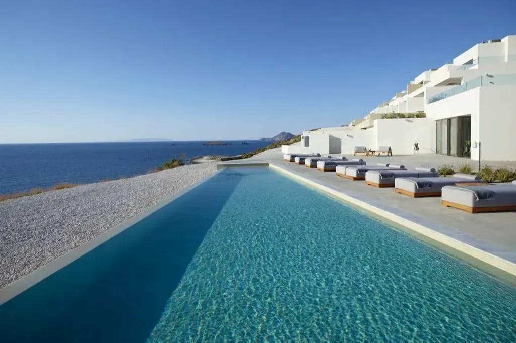 At Domes White Coast Milos, one of the coolest places to stay in Greece: a luxe and minimalist infinity pool area leads to Milos' rocky shoreline, with other Greek islands visible in the far distance