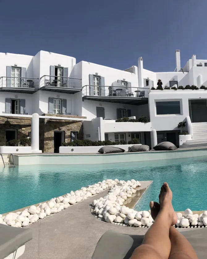 Joanne sun bathing on a lawn chair in front of a large white hotel and swimming pool surrounded by stones and palm trees