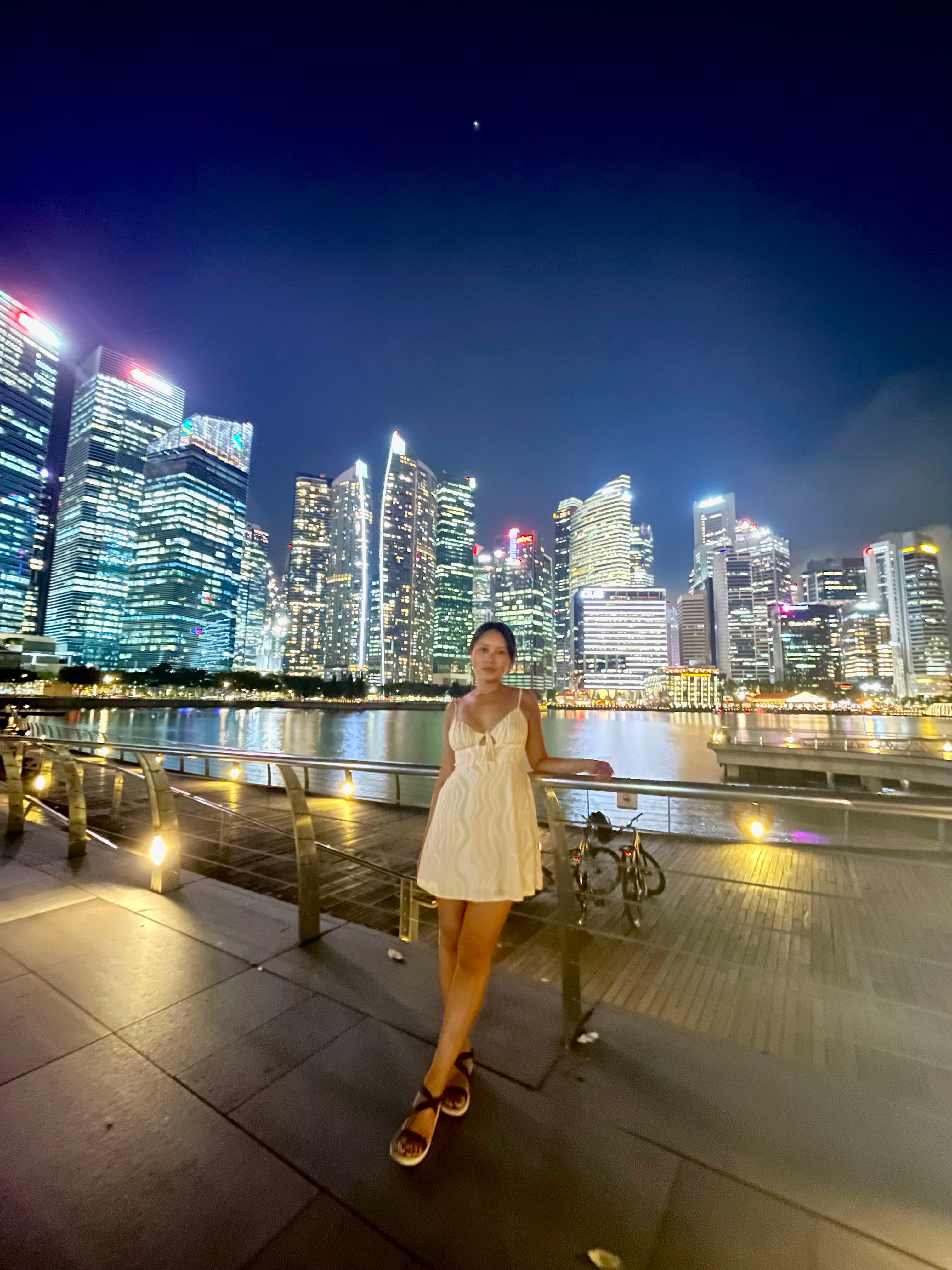Cam posing in a white dress in front of a city skyline at nighttime