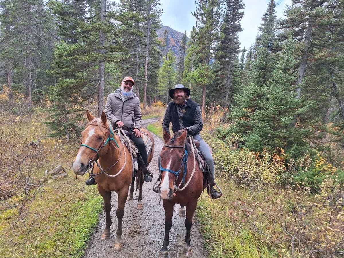 Two men on horse back on a trail among trees.