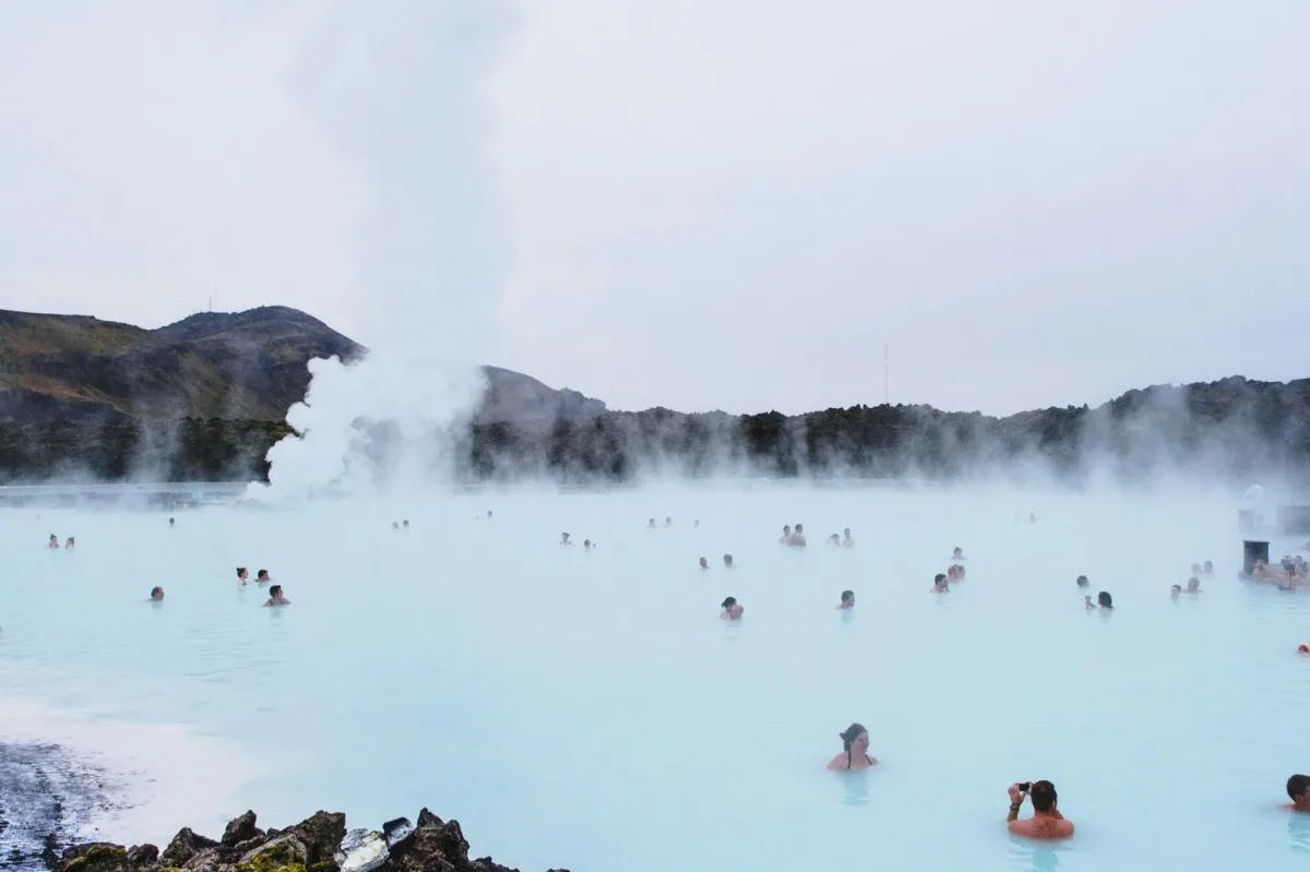 A few dozen travelers enjoy their time in Icelandic hot springs as steam rises up to cover mountains in the distance