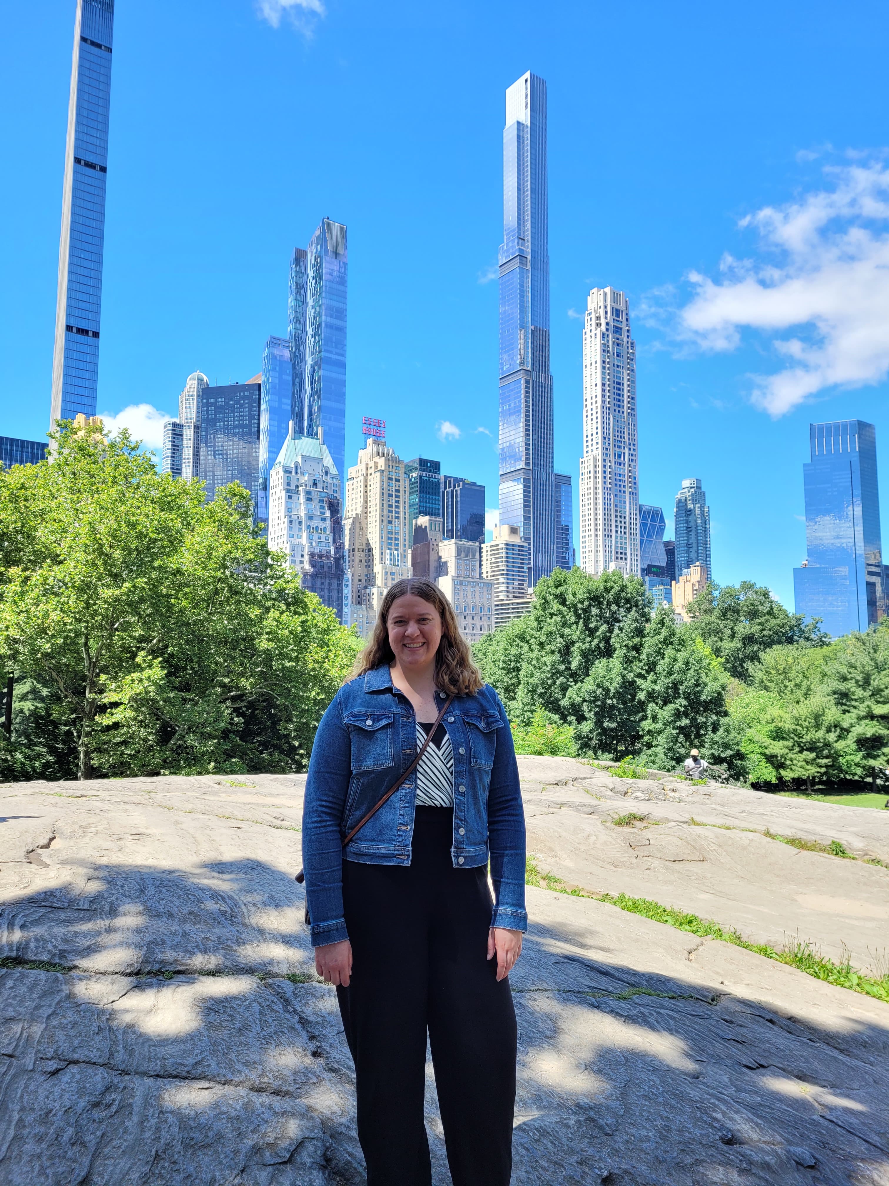 Stephanie standing on a boulder in front of trees and bushes with a backdrop of a city skyline.
