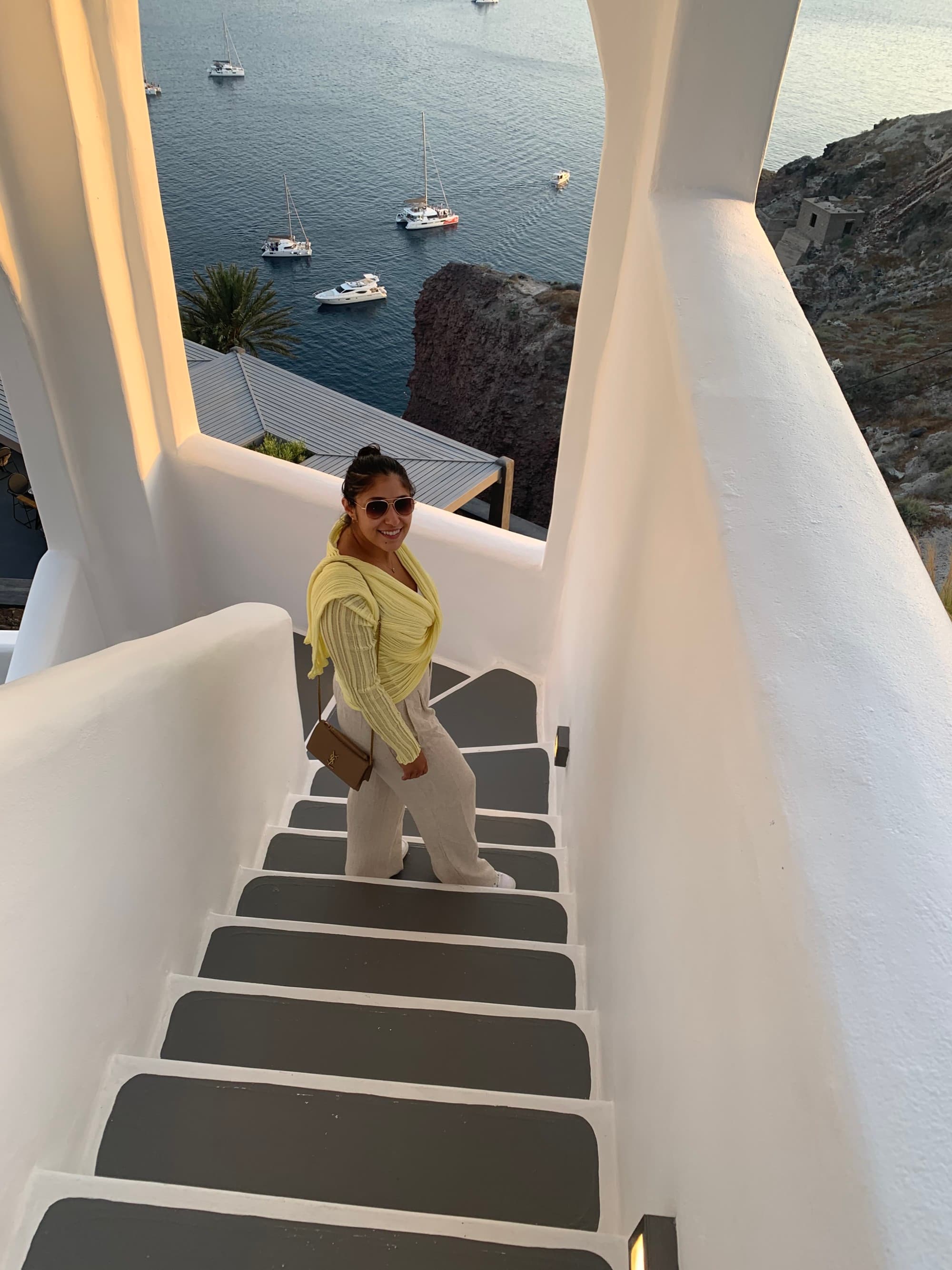 A woman in white pants, sunglasses, and a yellow top walks down a narrow stairwell overlooking the ocean.