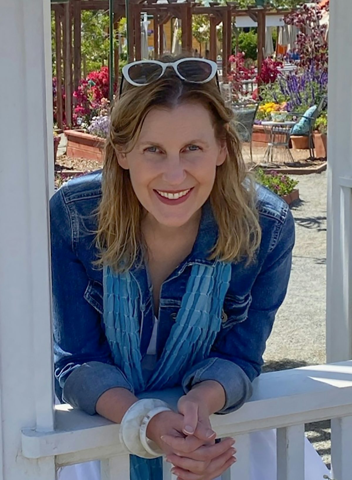 Fora travel agent Cara Todd wearing jean jacket, blue scarf and white sunglasses smiles with flowers in the background
