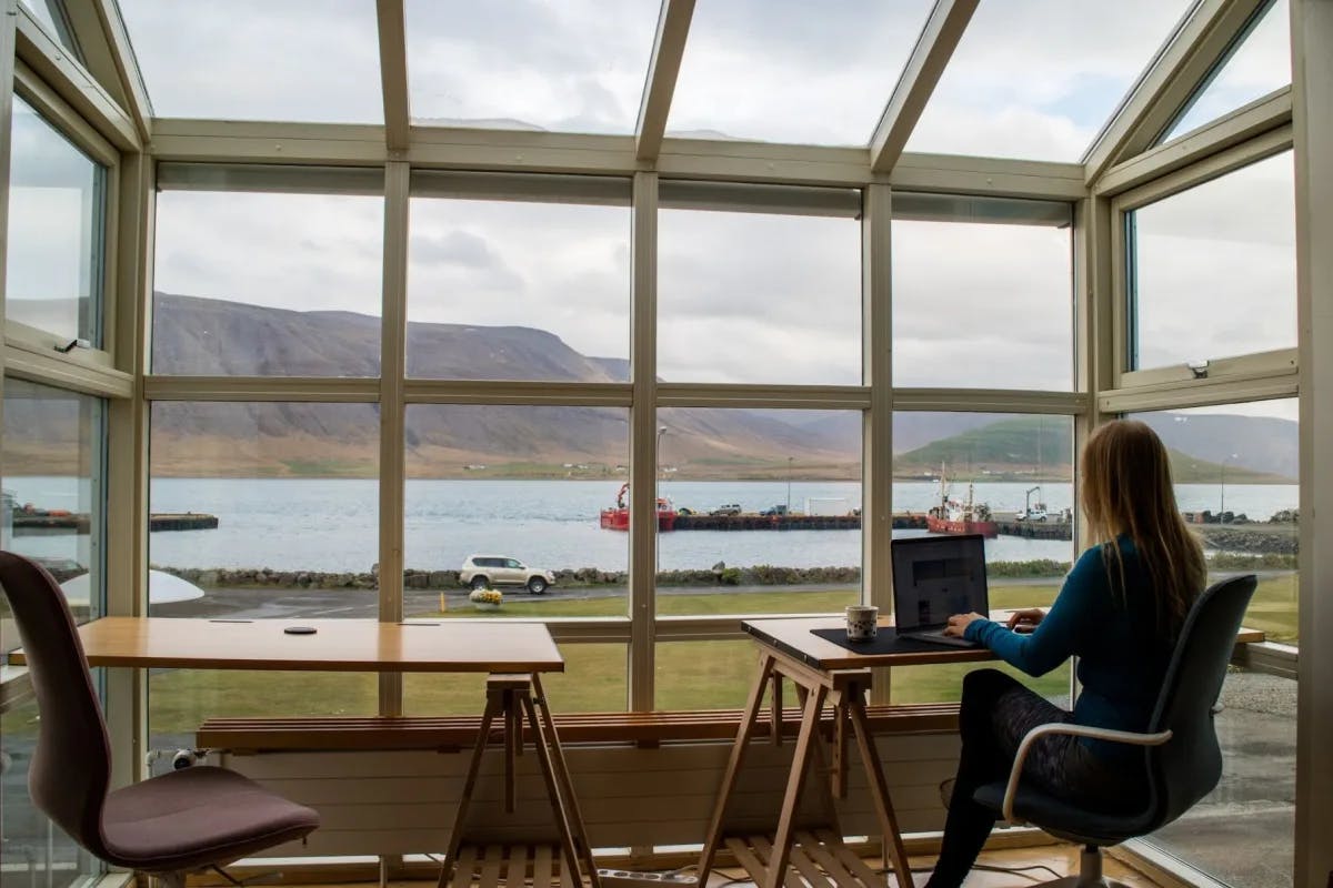A woman in trendy clothing works in the warmth of a coffeeshop with windows prominently reveal rural Iceland behind her