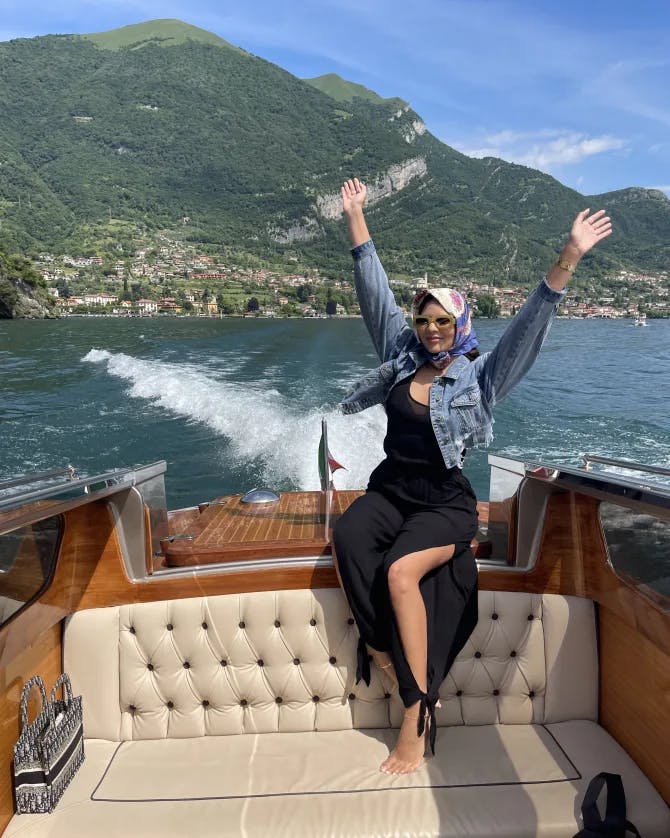 Picture of Aleah on a boat with her hands raised above her head and a green mountain in the background against the rippling blue water