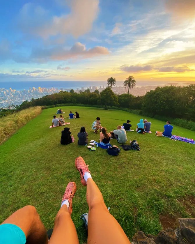 sunset photo on top of a hill with people sitting in a grassy area for relaxation after a hike. Palm trees and a city below in the distance. 