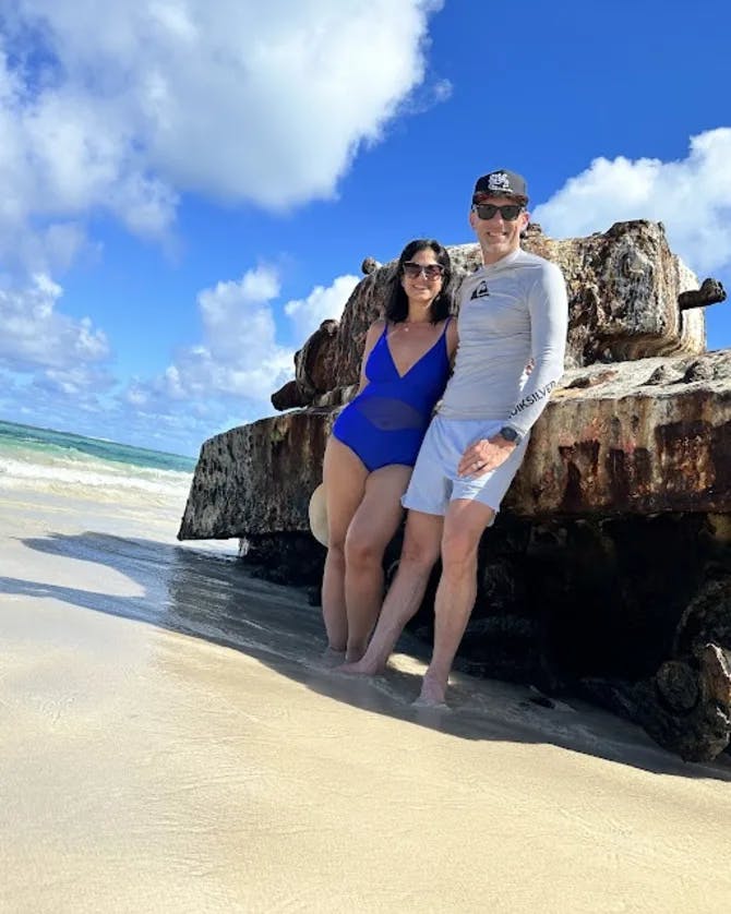 Mariela wearing a blue swimsuit while standing next to a man on the beach leaning against a large piece of machinery