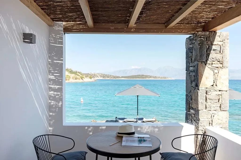 A snippet of Crete's shoreline and the sea is visible through a chill bungalow-style seating area at Minos Beach Art Hotel