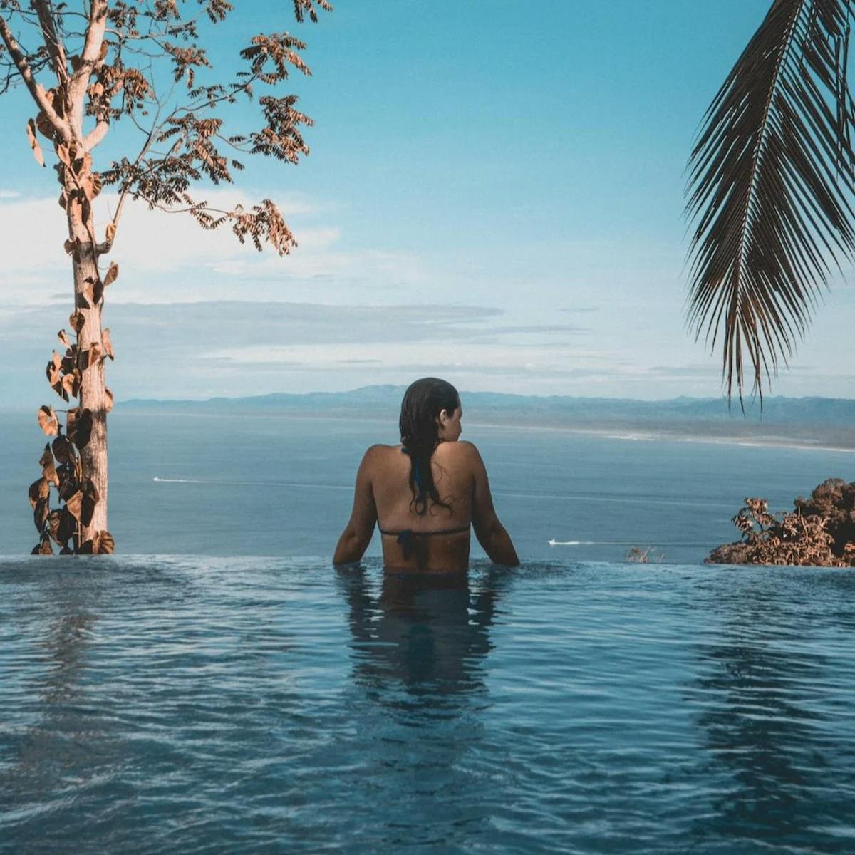 A woman in an infinity pool leans against the edge, taking in a wide view of the far off tropical coast of a bay.